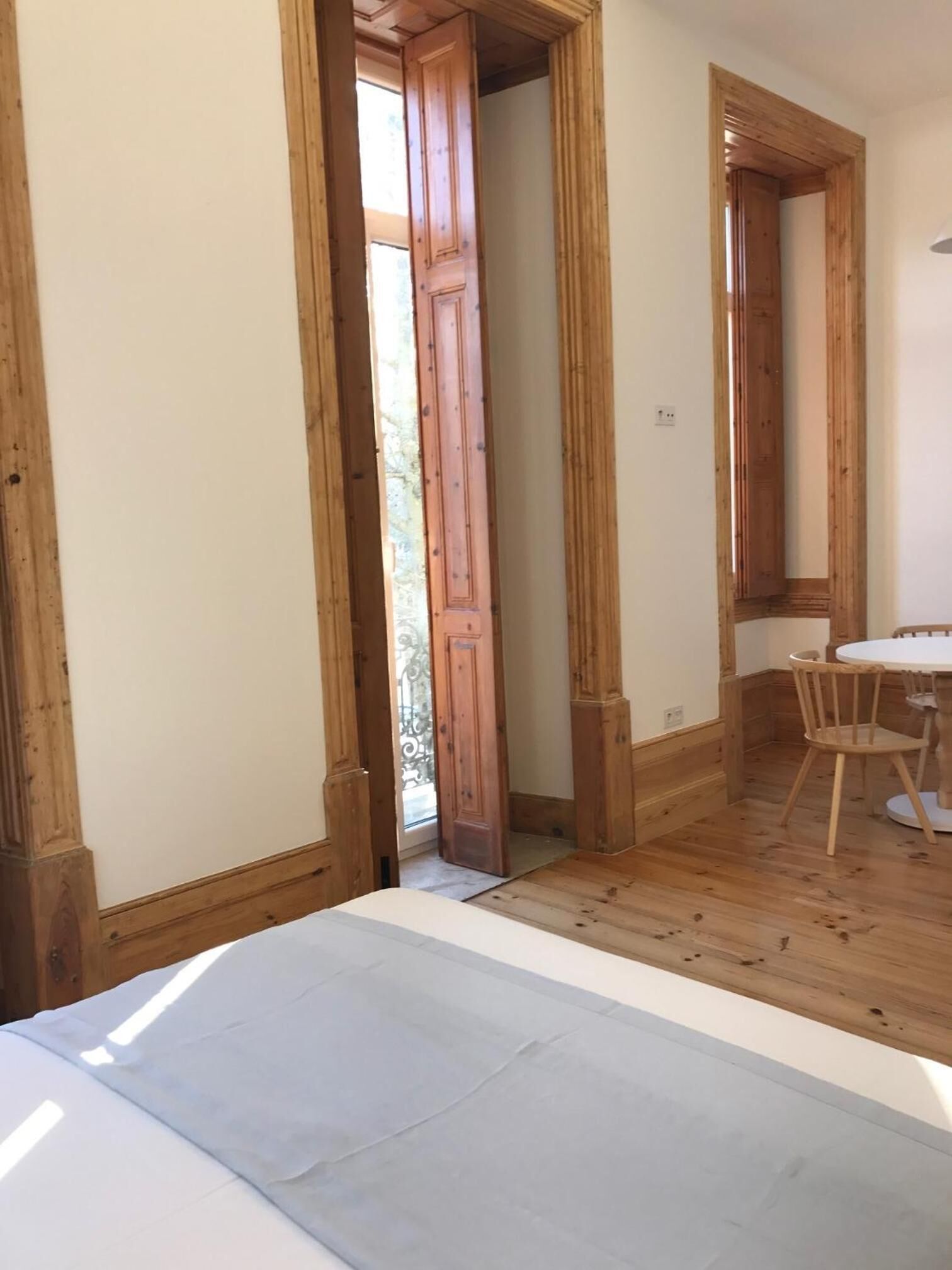 Una habitación con paredes y suelos de madera, una cama blanca, una pequeña mesa con sillas y una puerta que da a un balcón.