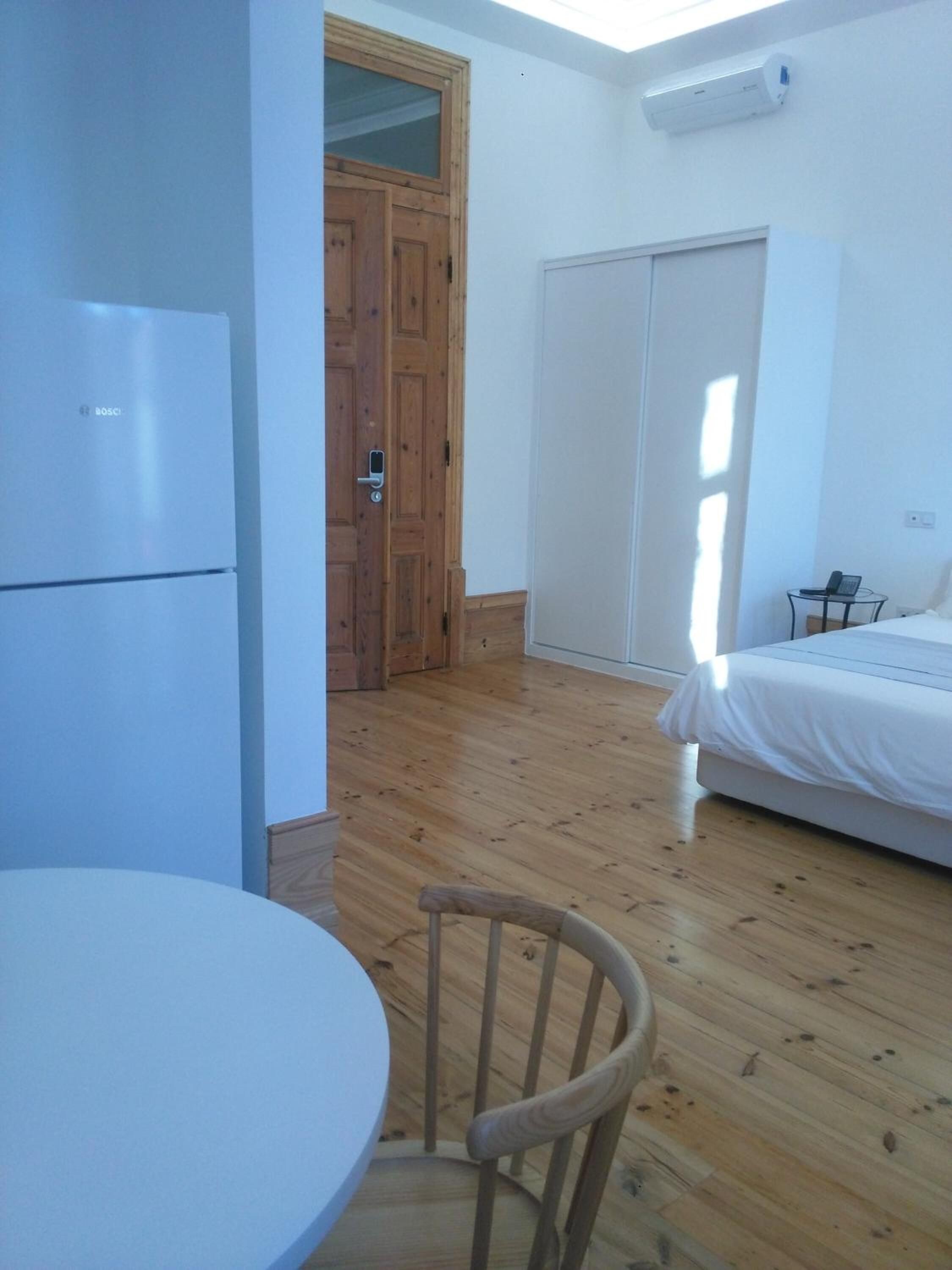 Una habitación con piso de madera, una cama, una mesa blanca y una silla. Hay una puerta con ventana y un armario blanco.