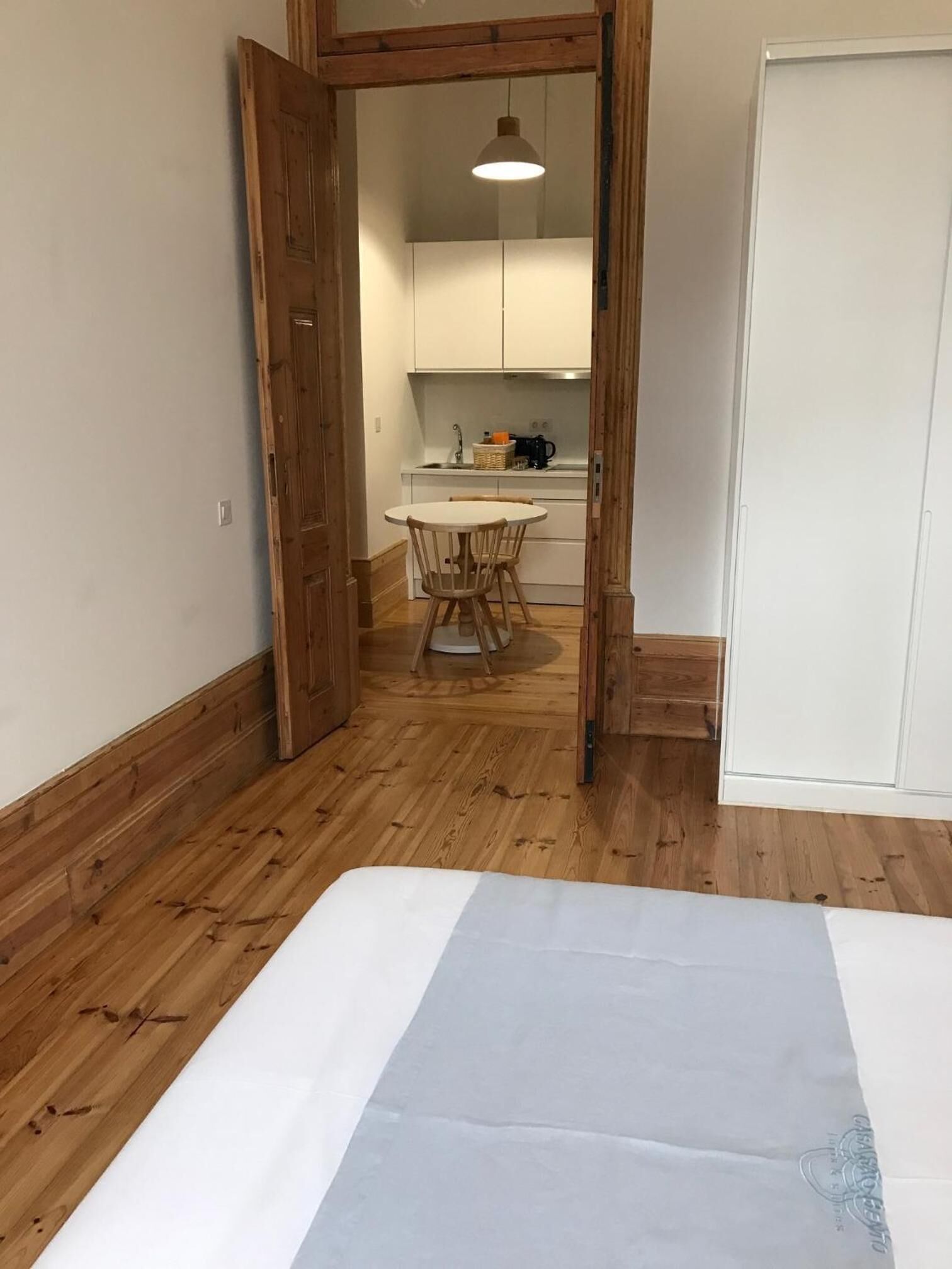 A room with a bed, wooden flooring, a small table with chairs, a kitchen area with cabinets, and a hanging light fixture.