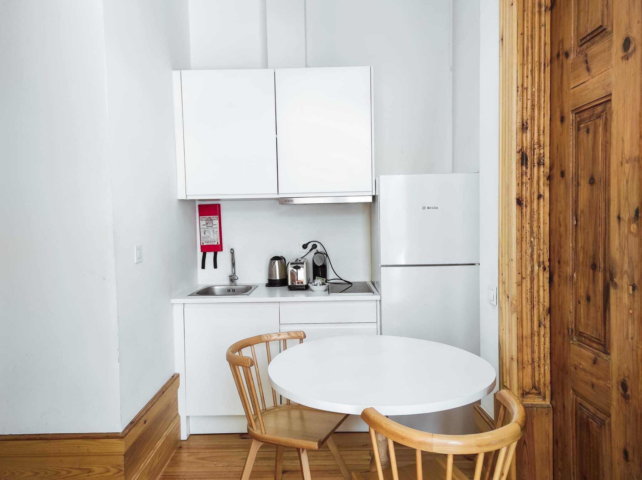Una pequeña kitchenette con armarios blancos, un refrigerador Bosch, un lavamanos, una tetera, un tostador y una silla de madera.