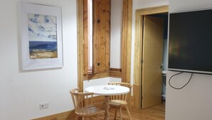 Een kamer met een houten vloer, een ronde tafel met stoelen, een schilderij aan de muur en een aan de muur bevestigde televisie.