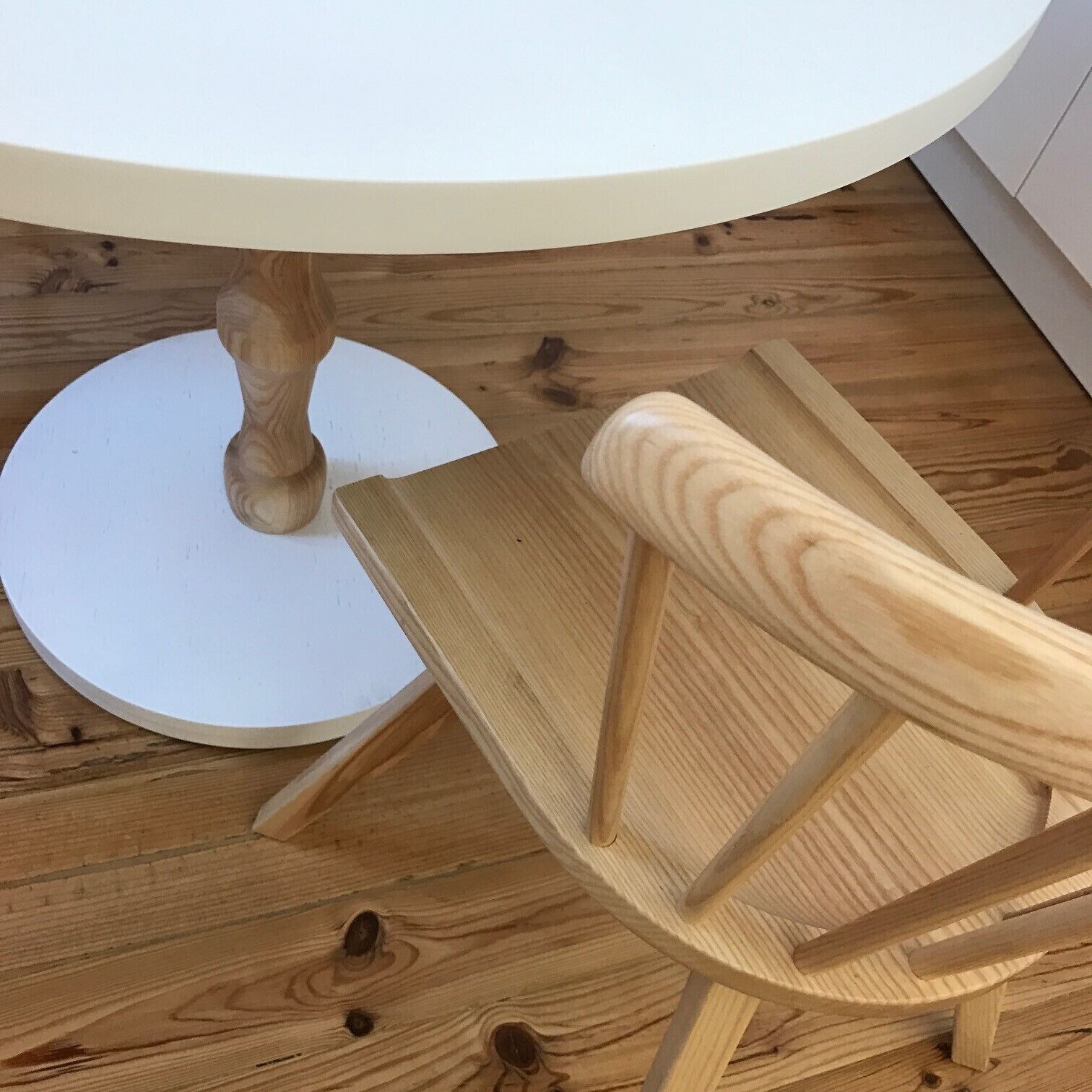 A round wooden table with a white top, a wooden chair with a curved backrest, and a wooden floor with visible grain patterns.