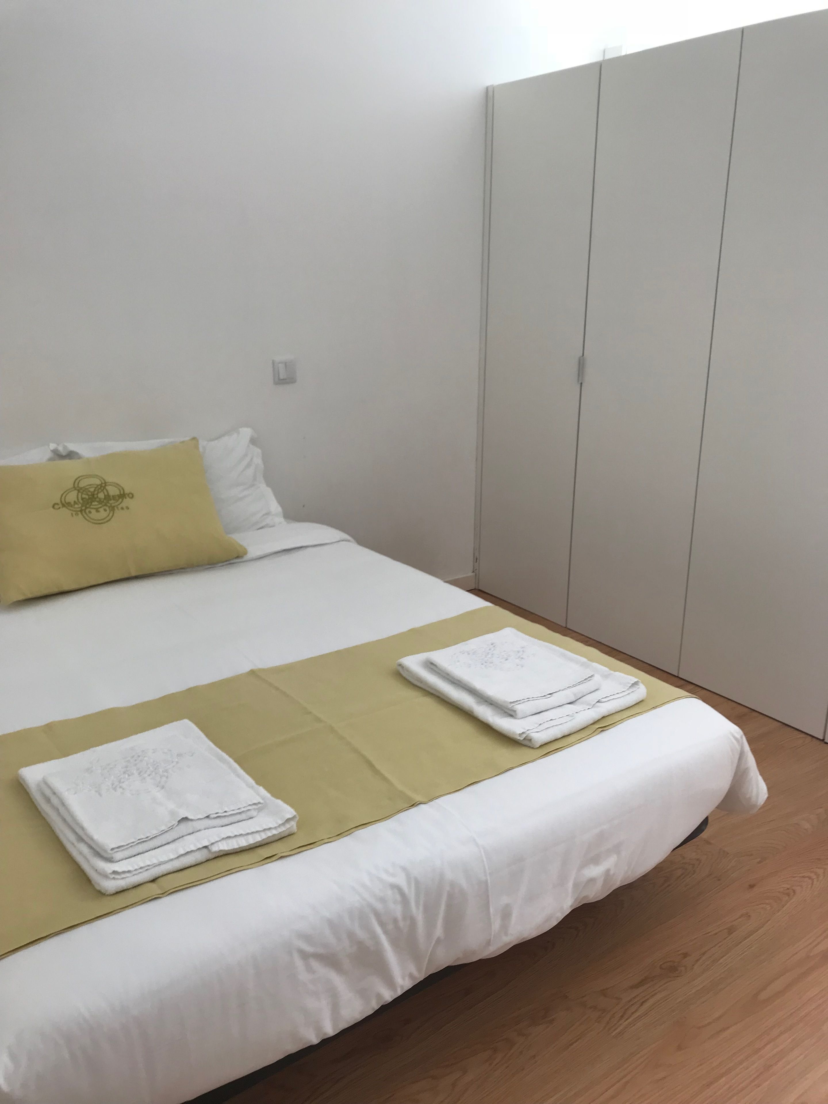Una cama bien hecha con sábanas blancas y un cojín amarillo decorativo, una toalla blanca doblada sobre la cama, y suelo de madera.