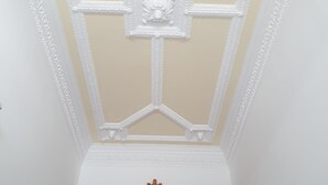 Een kamer met een hoge plafond, versierd met gedetailleerde witte sierlijsten en een centraal decoratief element. Daaronder is een houten, gewelfde deuropening met een glas-in-loodraam zichtbaar.