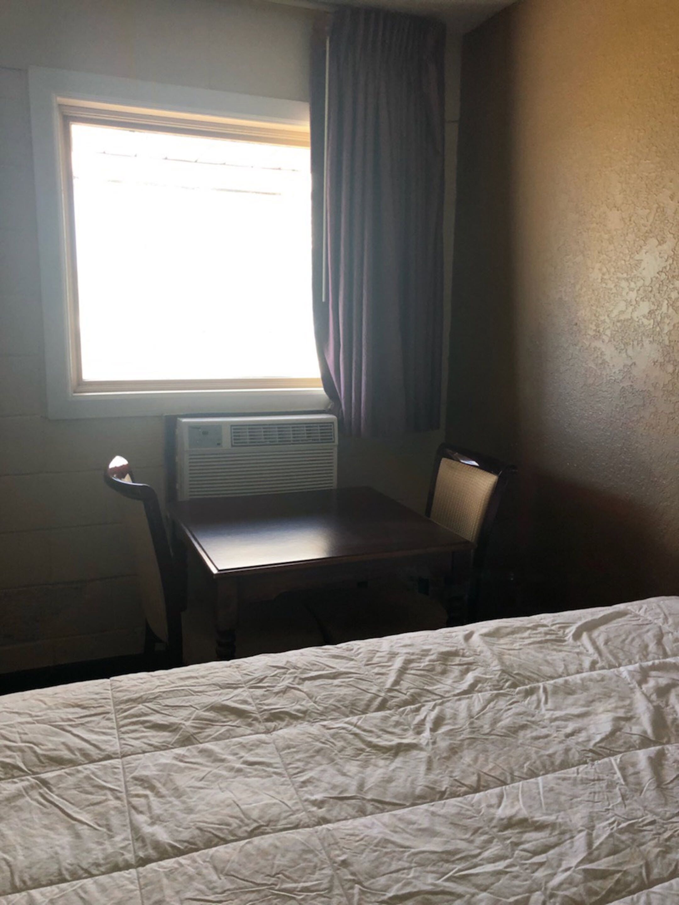 Habitación de hotel con cama, una mesita, una silla, ventana con cortinas y un equipo de aire acondicionado.