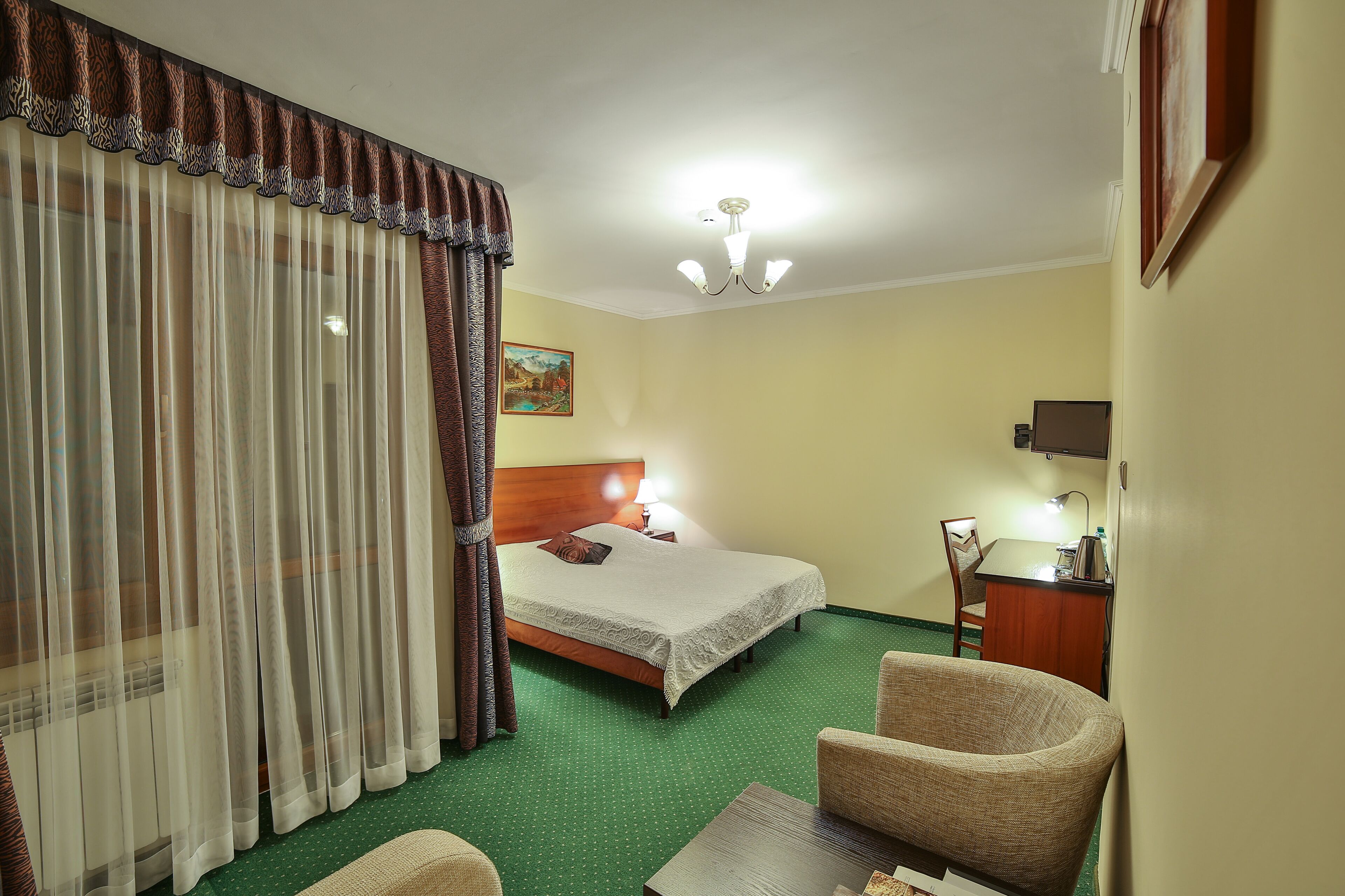 A hotel room with a bed, a desk, a chair, a lamp, and a painting on the wall.