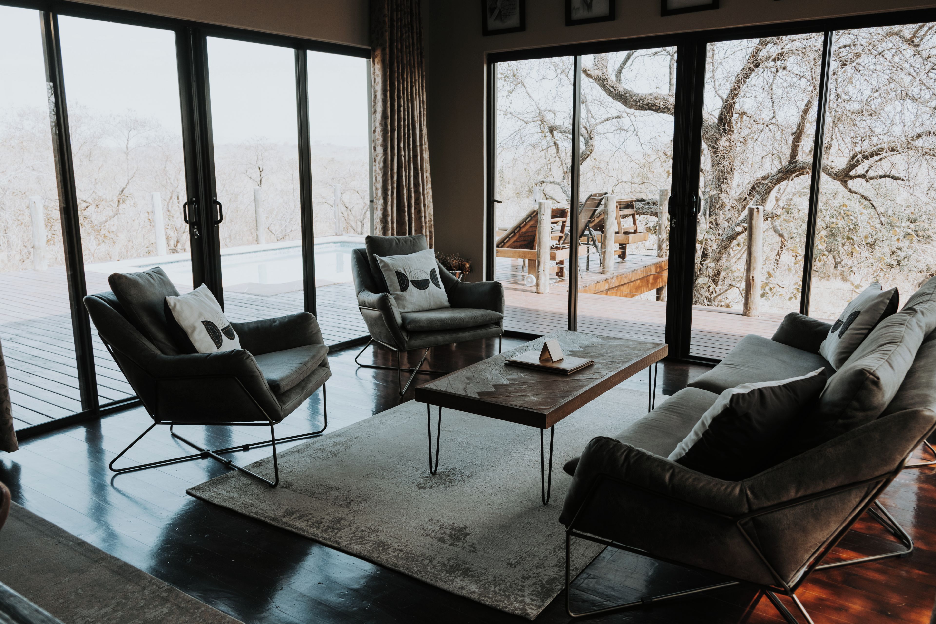 Ein modernes Wohnzimmer mit einer Sofagarnitur, einem Couchtisch und Blick auf eine Holzterrasse und Bäume durch große Fenster.