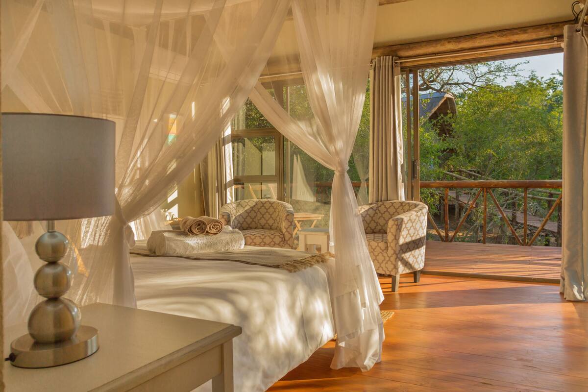 A bedroom with a canopy bed, a bedside table with a lamp, and a view of the outdoors.