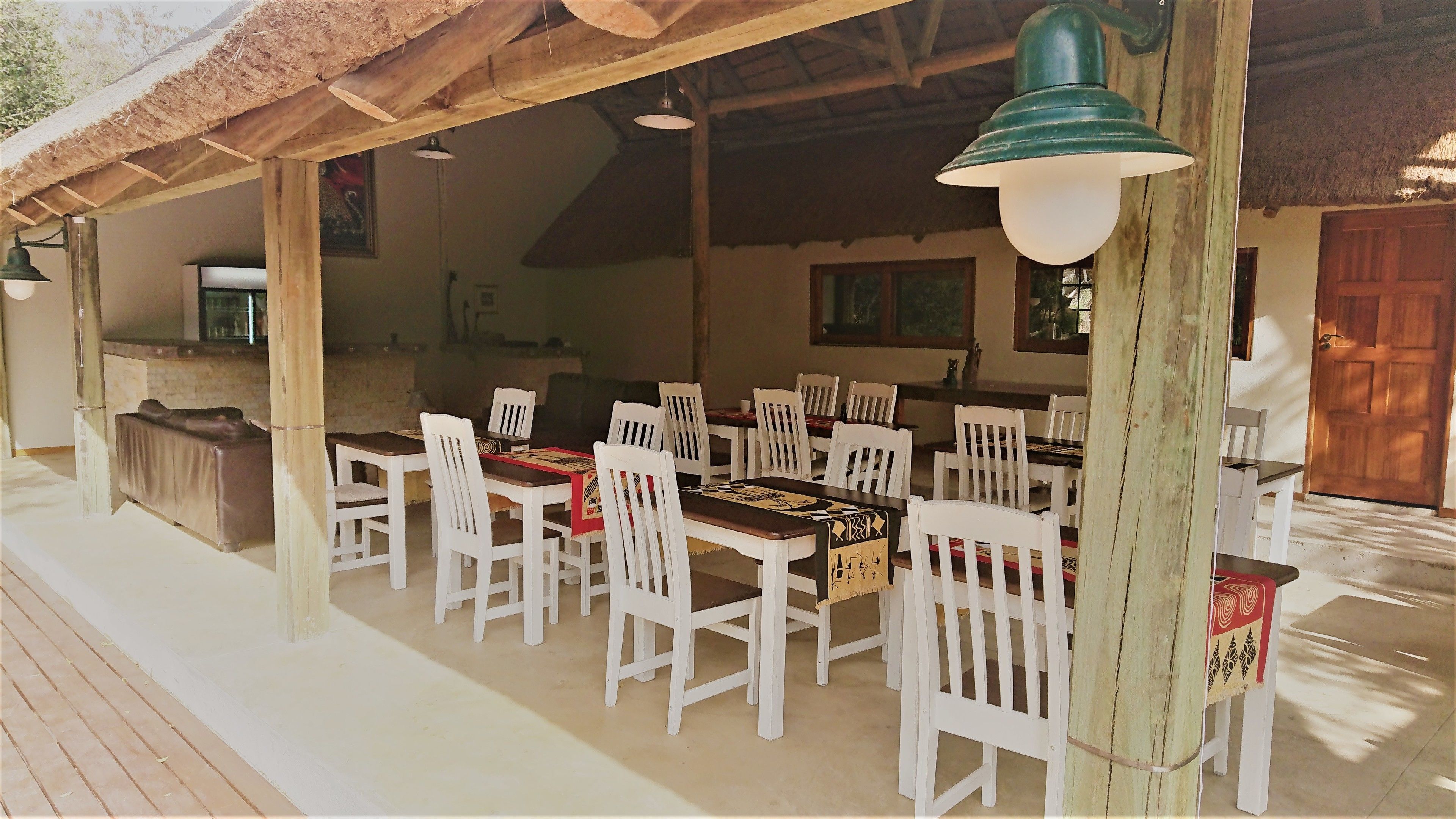 A covered outdoor dining area with white wooden tables and chairs, a thatched roof, and hanging pendant lights.