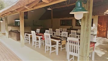 A covered outdoor dining area with white wooden tables and chairs, a thatched roof, and hanging pendant lights.