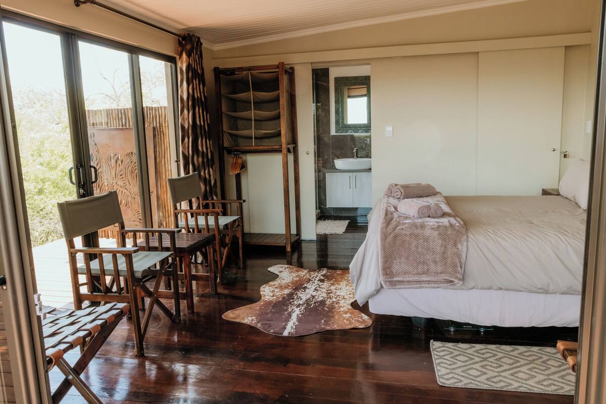 A bedroom with a bed, a wooden chair, a bench, and a view of the outdoors through a sliding door.
