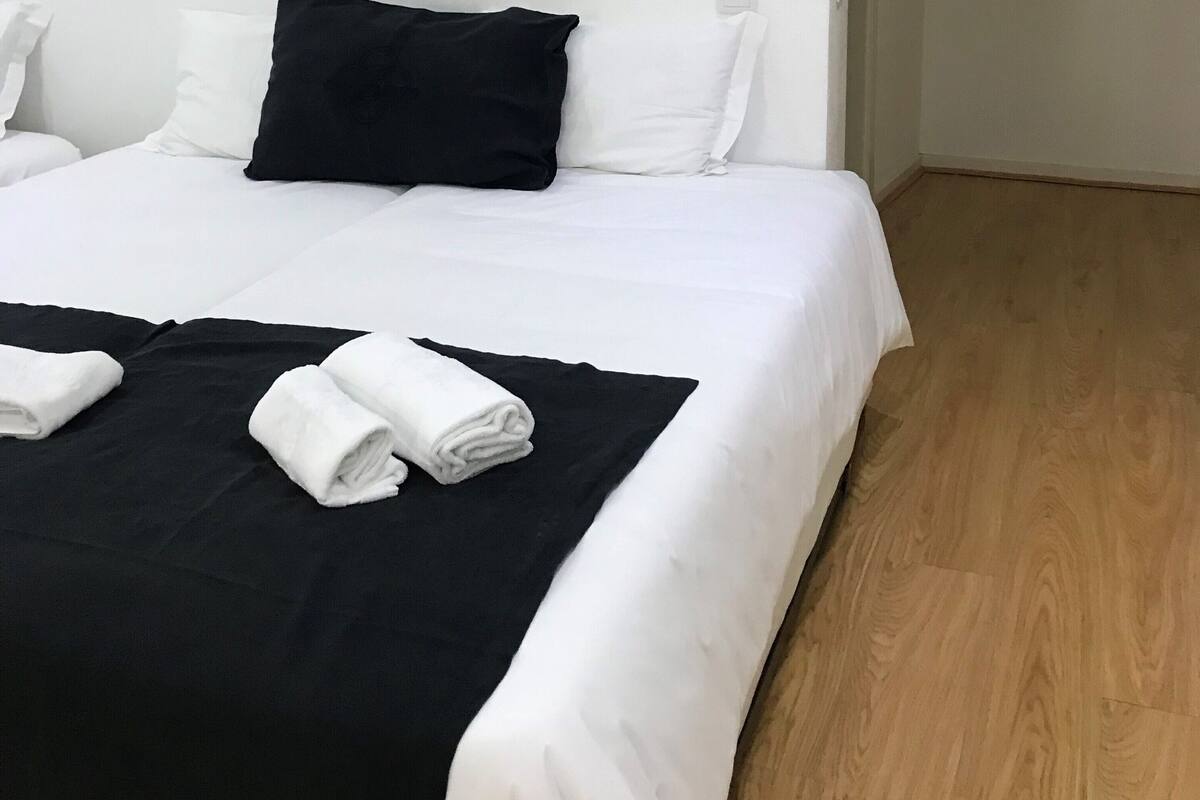 A neatly made bed with white and black bedding, a black pillow, and two rolled towels on a black bed runner.
