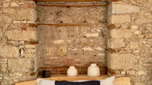 Una stanza con parete in pietra, travi in legno, un letto con biancheria bianca e due vasi bianchi su uno scaffale.