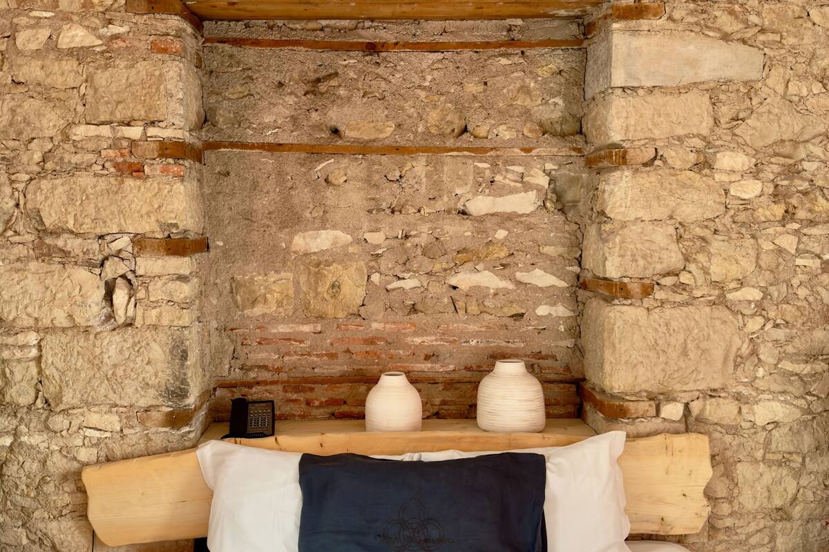 A room with a stone wall, wooden beams, a bed with white bedding, and two white vases on a shelf.