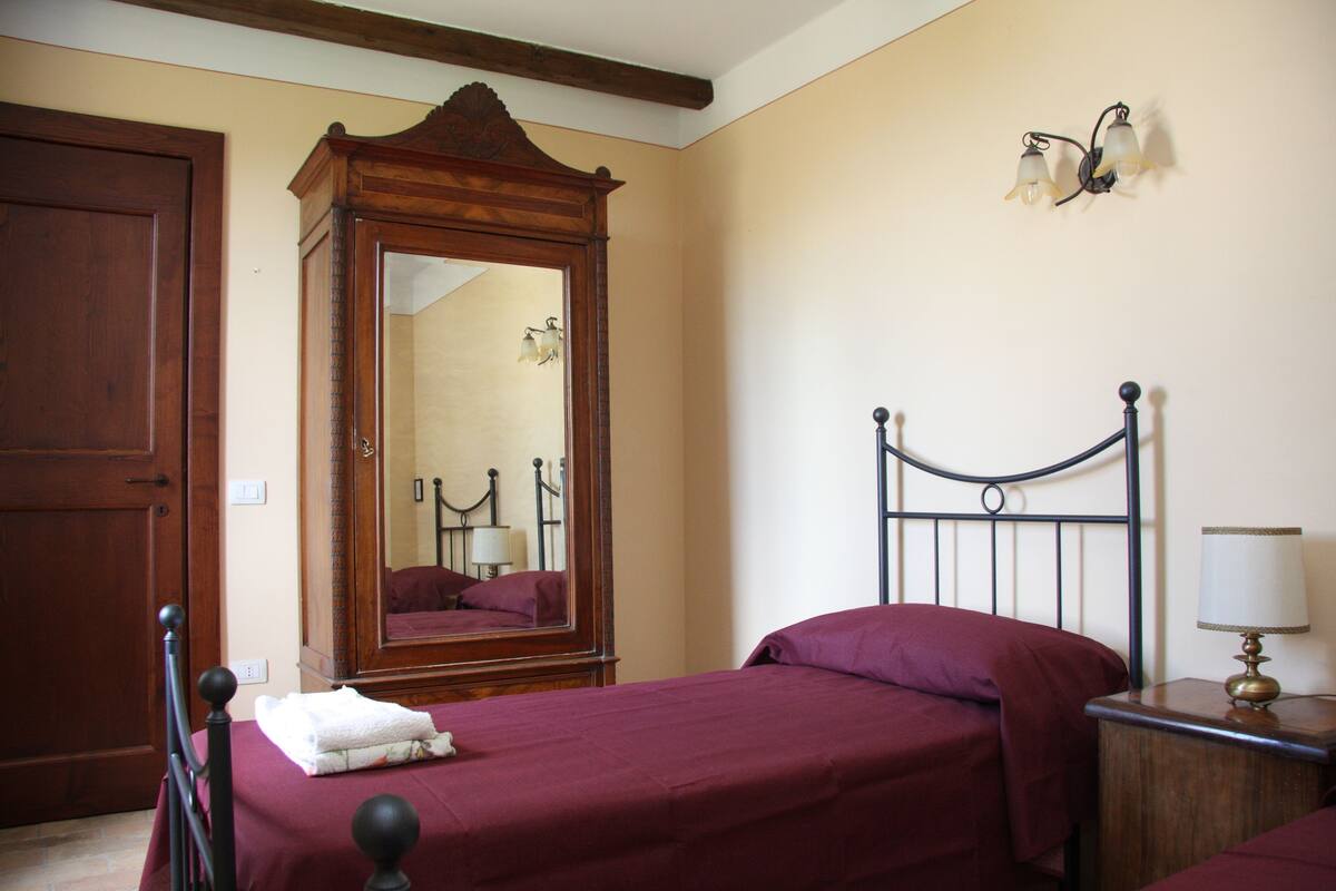 A bedroom with a wooden wardrobe, a bed with a headboard, a nightstand with a lamp, and a wall-mounted light fixture.