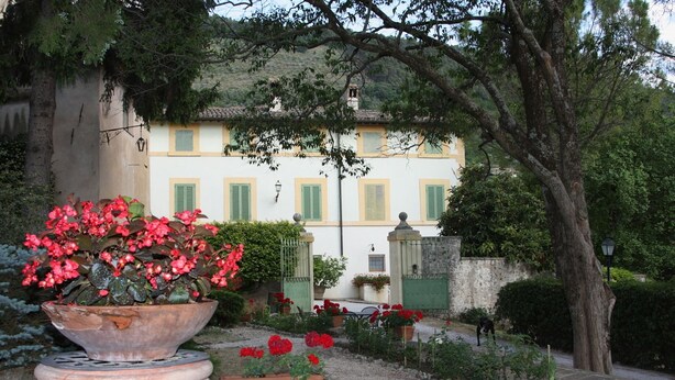 A garden with a large planter of red flowers, a white building with green shutters, and a stone wall. - Villa del Cardinale (Spoleto)