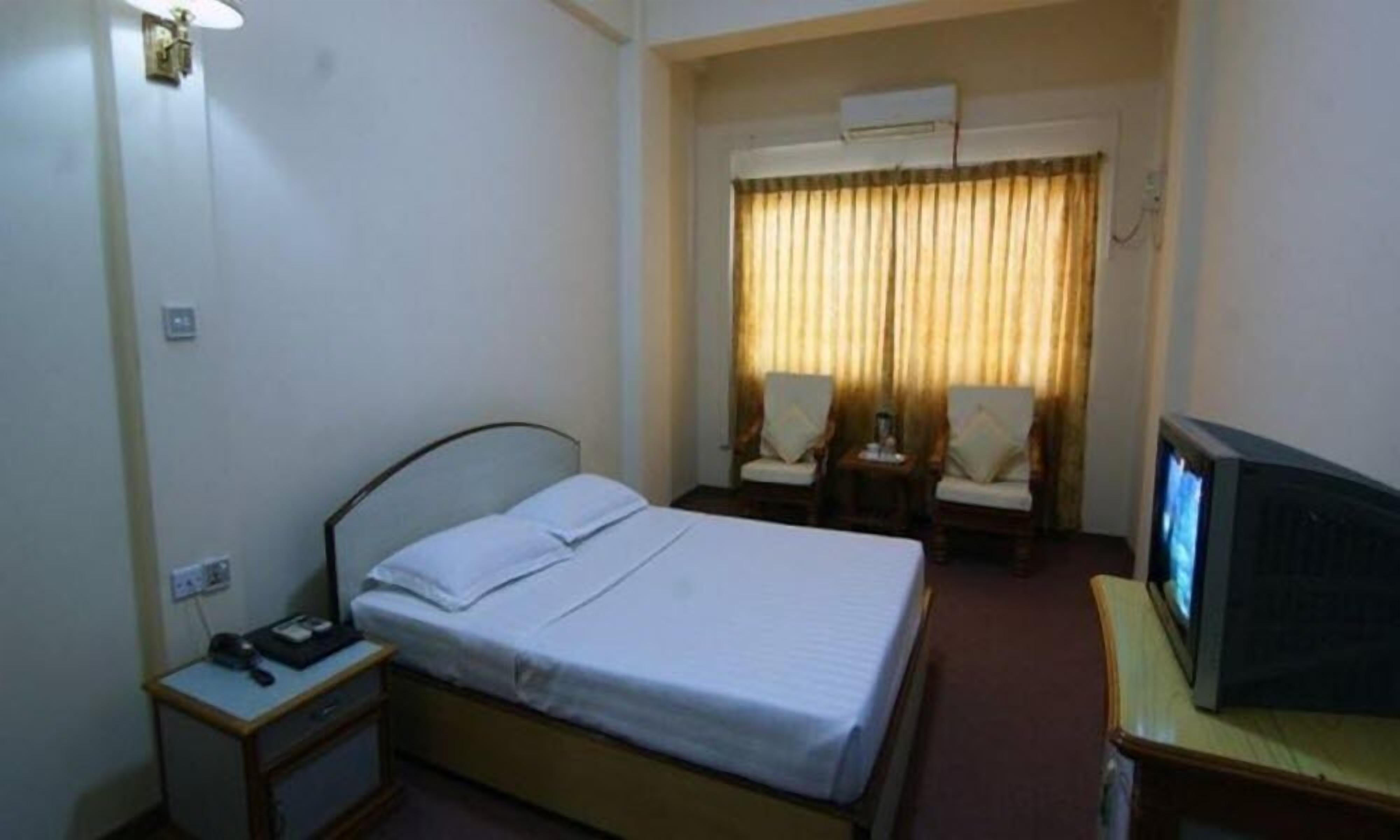 Hotellihuone, jossa on yksi sänky, yöpöytä lampun kanssa, tuoli, televisio ja ilmastointilaite.