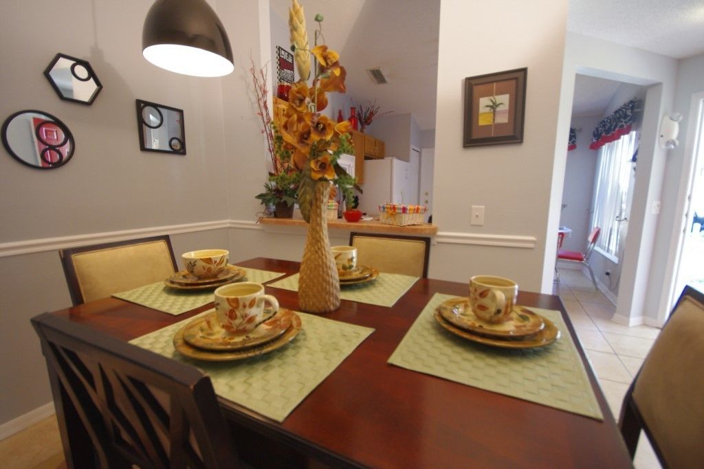 Uma área de refeições com uma mesa de madeira preparada para quatro pessoas, rodeada por cadeiras. Há um vaso decorativo com girassóis sobre a mesa, e as paredes são adornadas com quadros.
