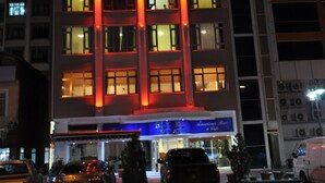 Een meerhoog hotel met verlichte rode en witte lampen op de gevel, een parkeerplaats met meerdere auto's, en een bord waarop 'American Bar & Grill' staat.