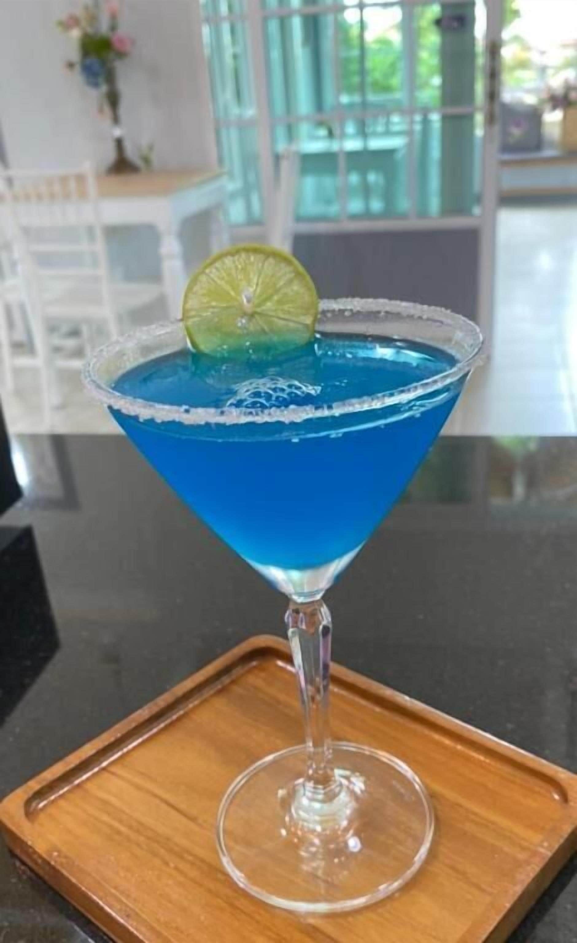 A blue cocktail in a martini glass with a lime garnish on a wooden tray.