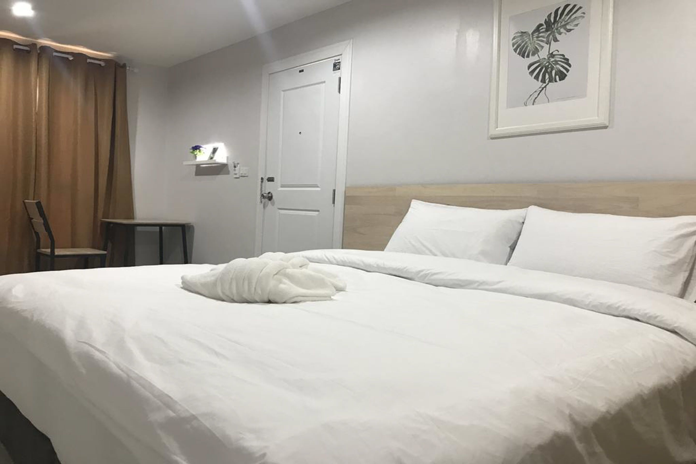 A neatly made bed with white linens and a folded towel, a wooden headboard, a white door, and a framed plant on the wall.