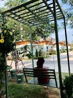 Un columpio con un banco debajo, rodeado de follaje y una zona residencial al fondo.