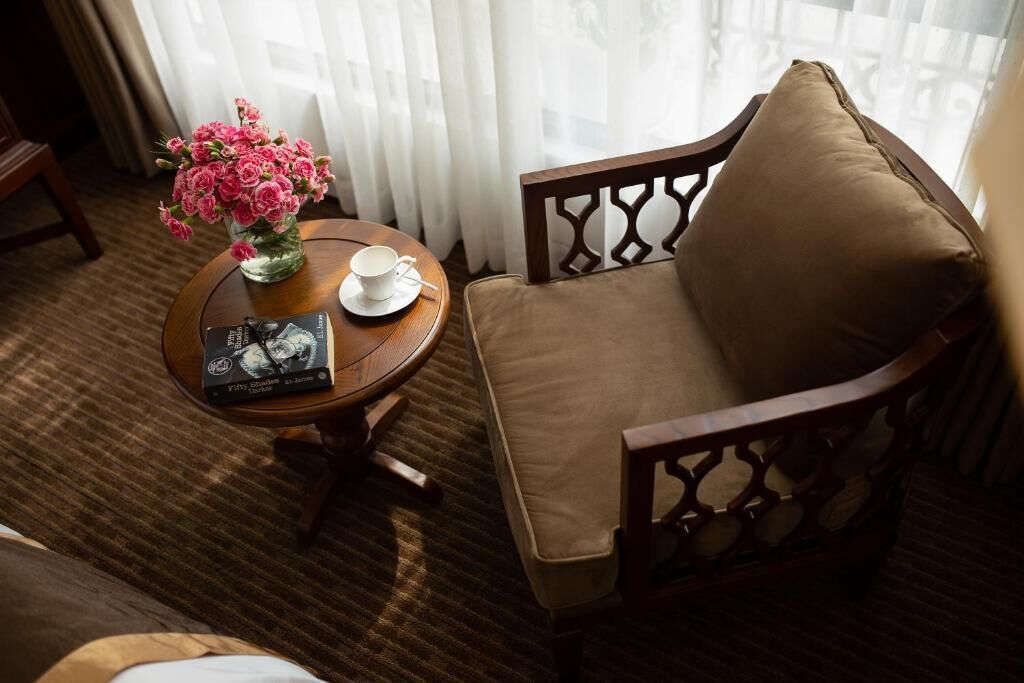 Ein Hotelzimmer mit braunem Sessel, einem kleinen runden Tisch mit einer Vase voller rosa Blumen, einer Tasse und einer Zeitschrift.