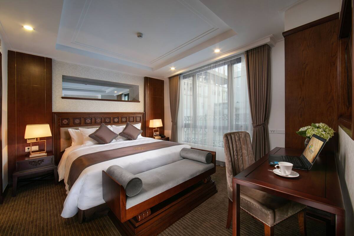A hotel room with a large bed, bedside tables with lamps, a desk with a laptop, and a view of the city through large windows.