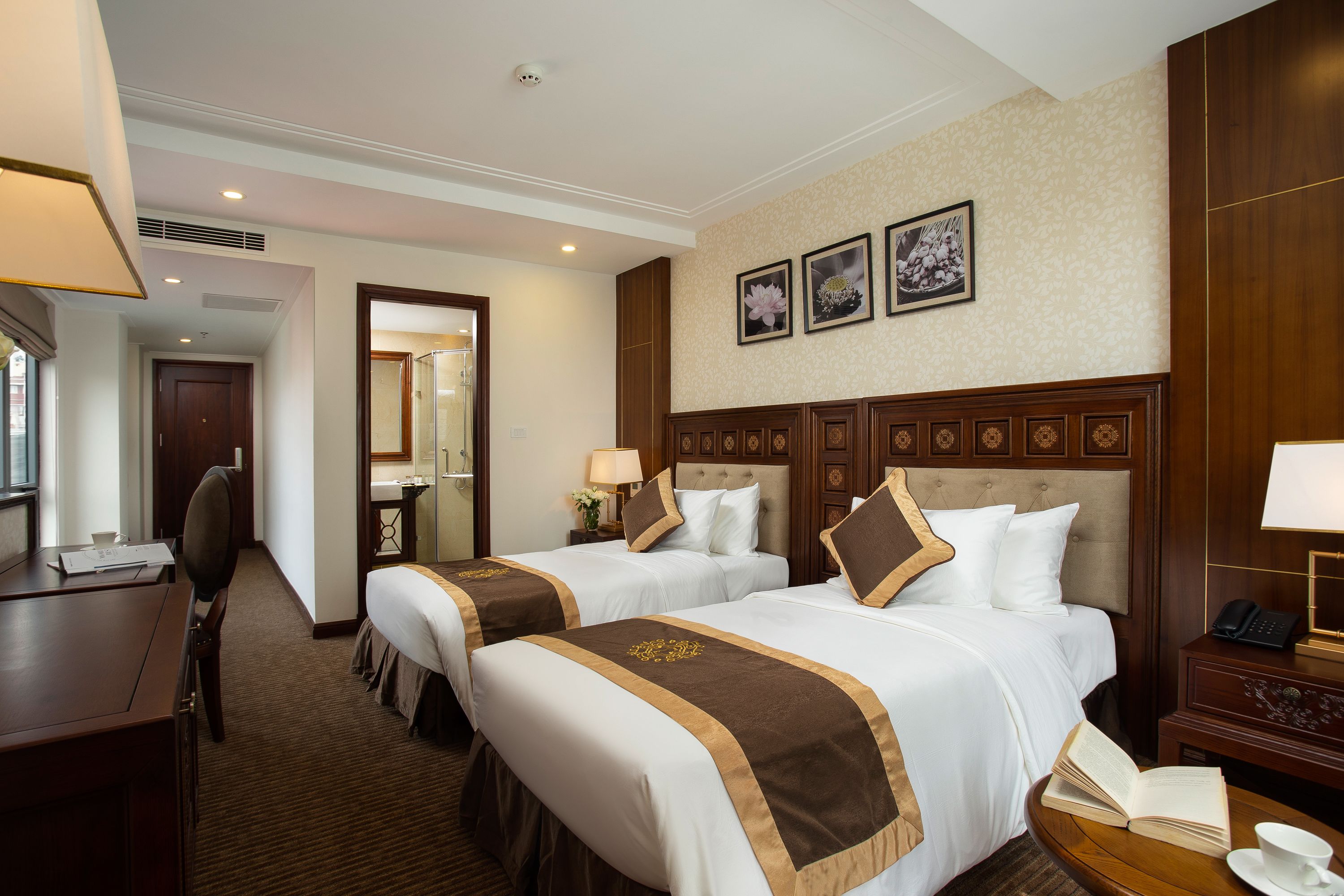 A hotel room with two beds, a desk, a chair, a bedside table with a lamp, and a telephone.