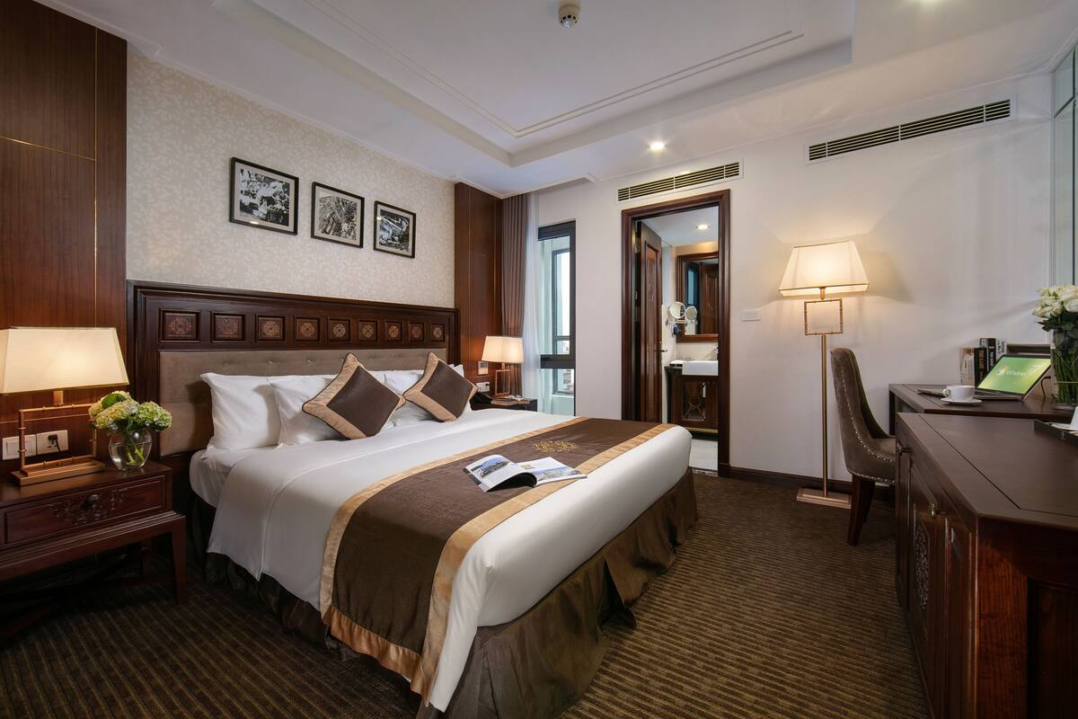 A hotel room with a large bed, a desk, a chair, a lamp, and a small table with a vase of flowers.