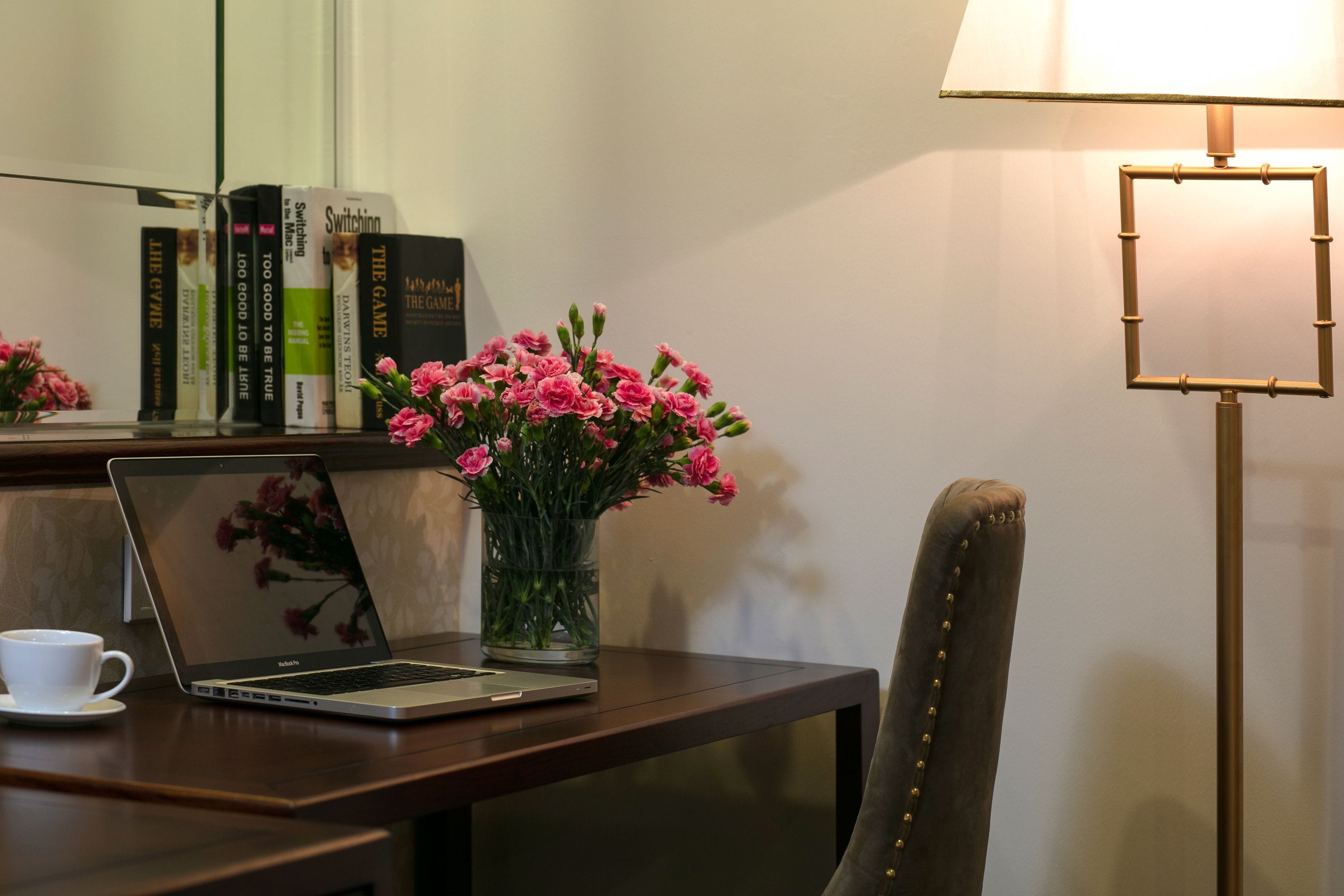 A laptop on a wooden table with a vase of pink flowers, a lamp, and books on a shelf.
