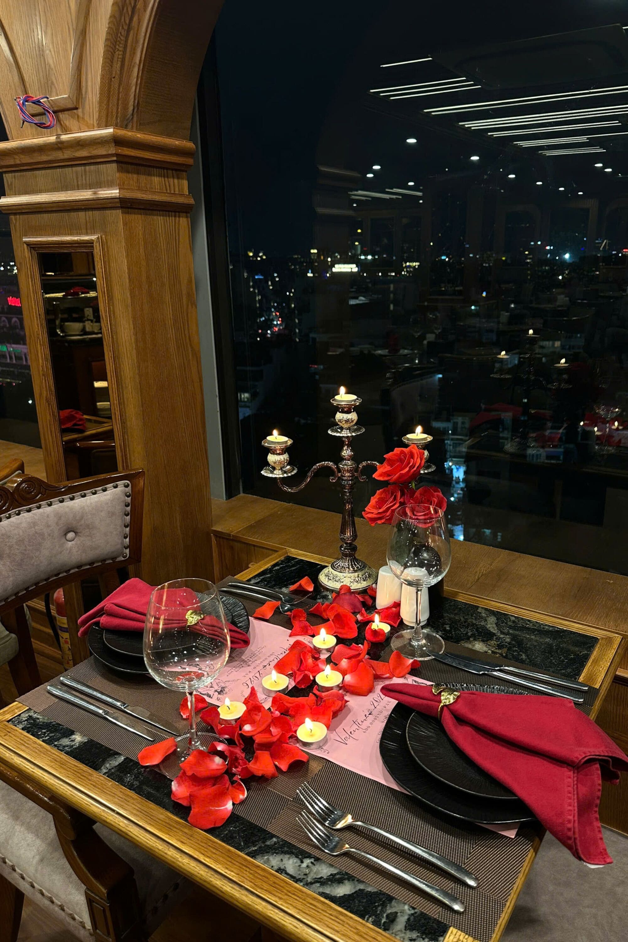 A dining table set with red roses, candles, and wine glasses, overlooking a cityscape.