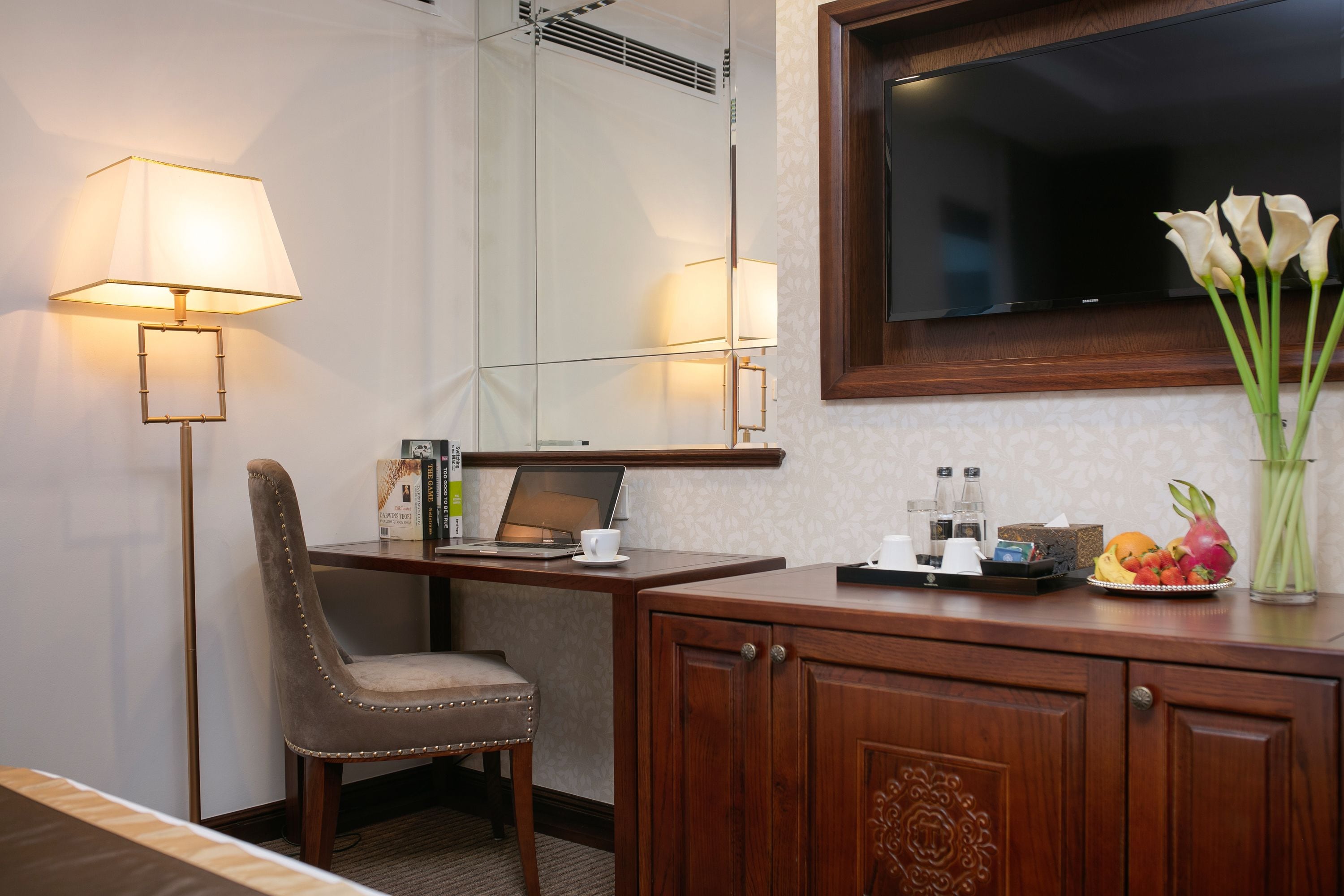 Een hotelkamer met een bureau, stoel, lamp, televisie en een kast met een bloemenvaas.