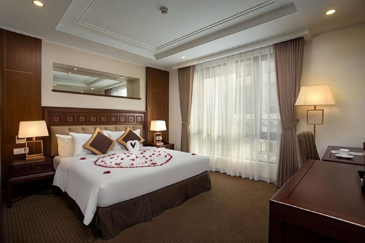A hotel room with a large bed, two bedside lamps, a desk, and a large window with curtains.