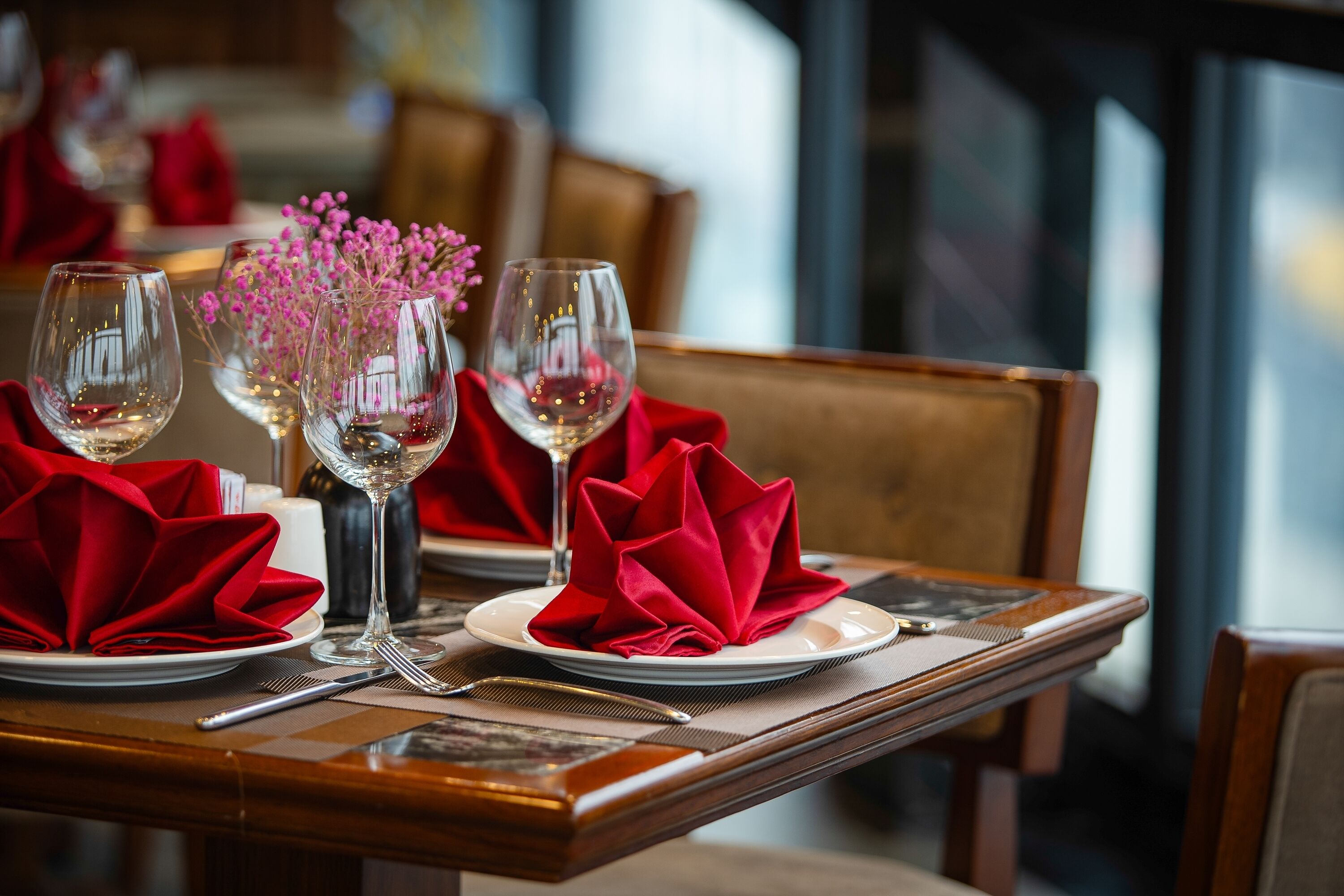 A dining table set with red napkins, clear wine glasses, and a vase with pink flowers.