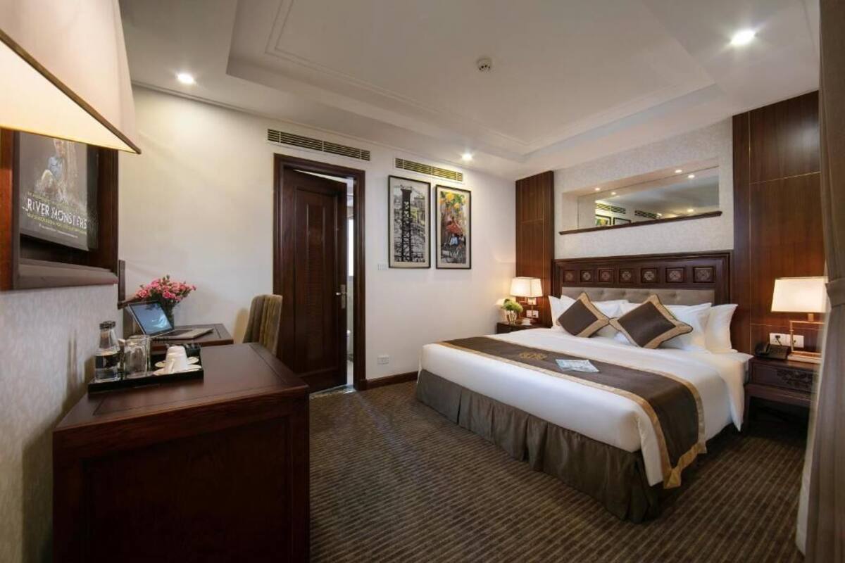 A hotel room with a large bed, a desk with a laptop, a mirror, and framed pictures on the wall.