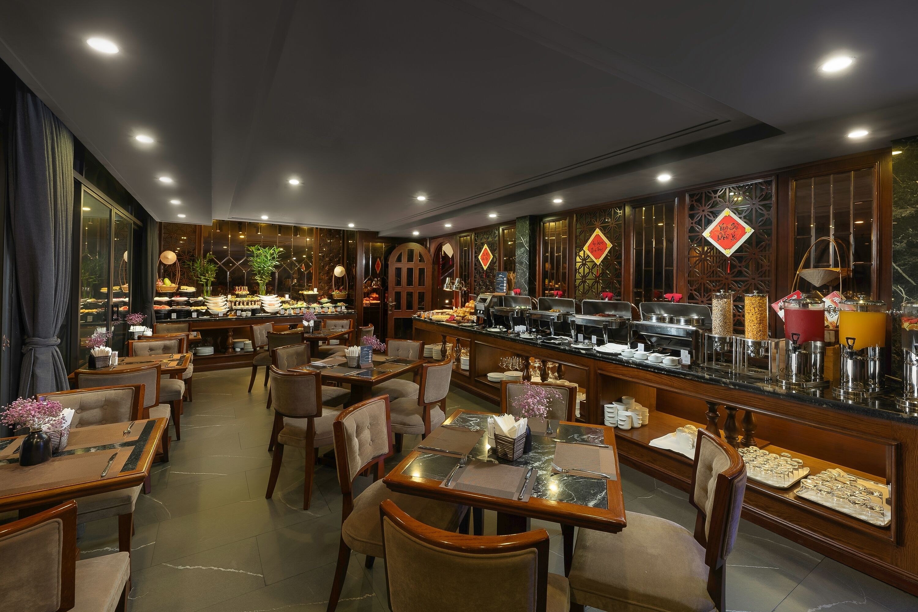Un restaurante con varias mesas y sillas, una zona de bufé y detalles decorativos como plantas y arte mural tradicional.