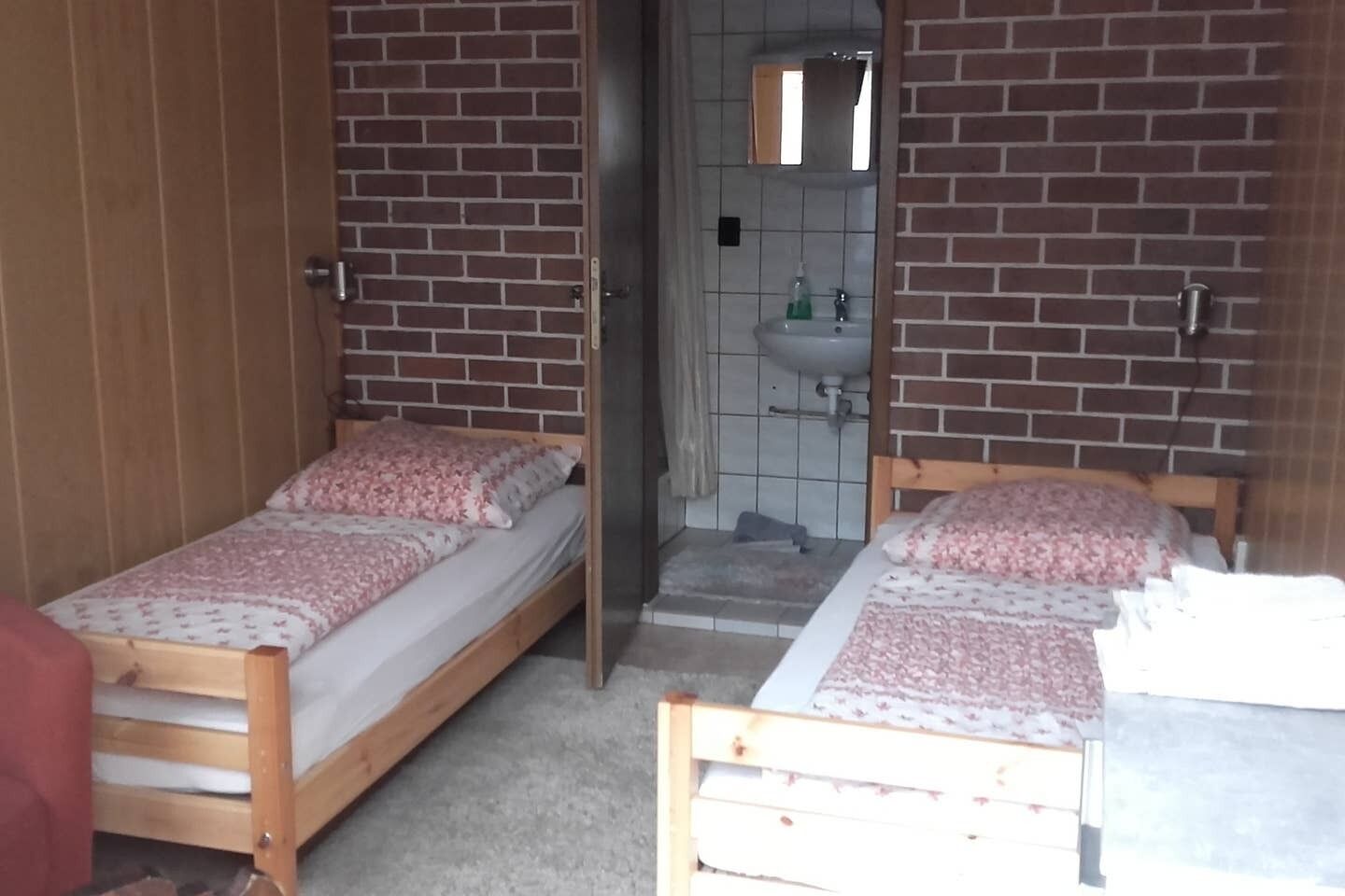 A small, compact room with two single beds, a wooden door, a small bathroom with a sink and mirror, and a tiled floor.
