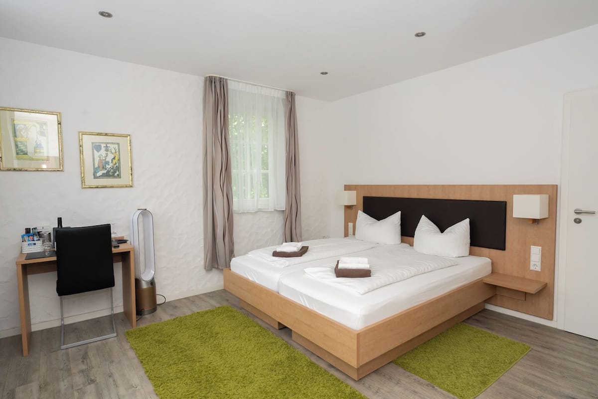 A modern hotel room with a wooden bed, a desk with a chair, a standing heater, and a green rug.