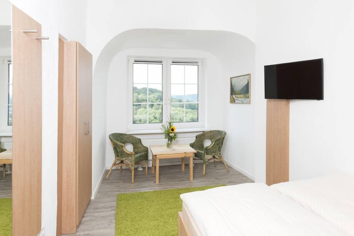 A bedroom with a bed, a TV, a window with a view, and a small table with chairs.