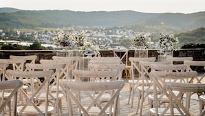 A terrace with rows of wooden chairs and tables, set against a backdrop of a town and hills.