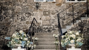 A stone building with a staircase leading to a second floor, white flower arrangements at the base, and a black railing.