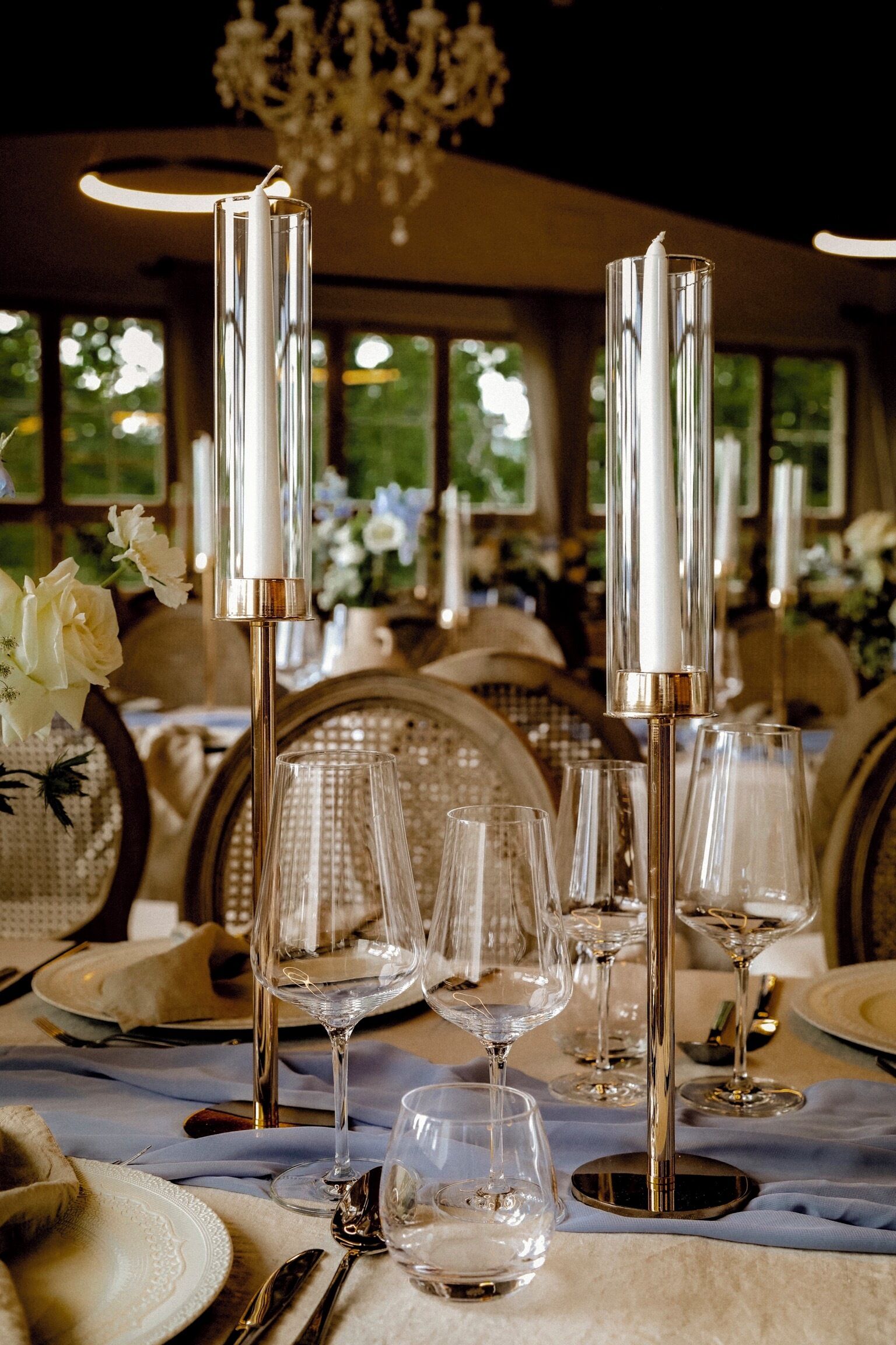 Una tavola imbandita con calici, piatti e posate, con alti candelabri e un lampadario sullo sfondo.