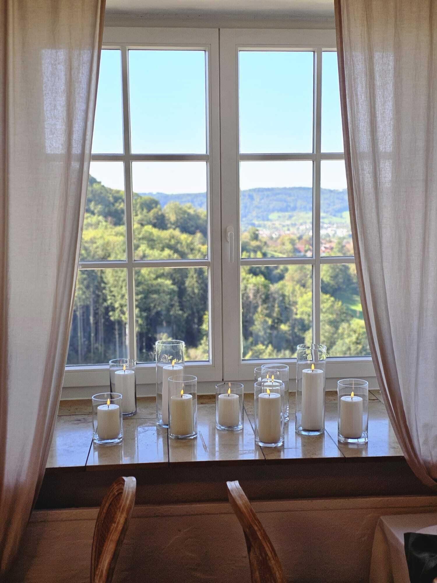 Una habitación con un ventanal amplio, sillas de madera y varias velas sobre un alféizar.