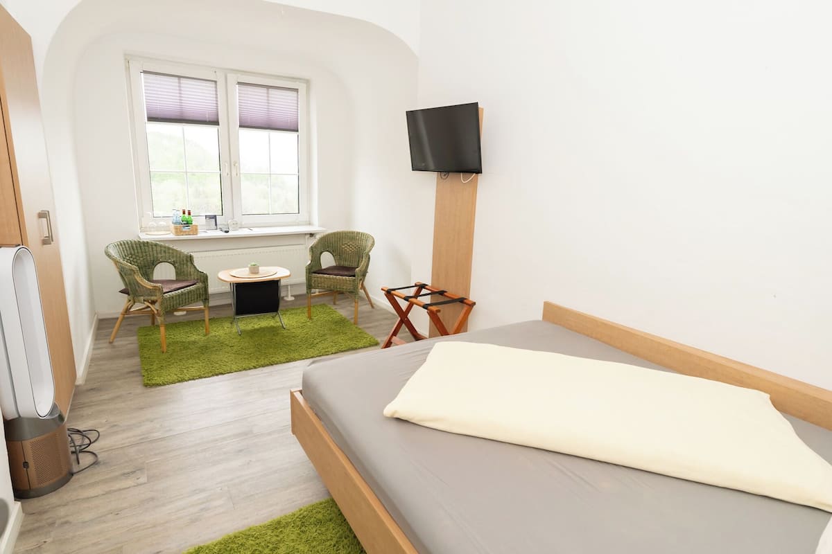A bedroom with a bed, a TV mounted on the wall, a window with blinds, two wicker chairs, a small table, and a green rug.
