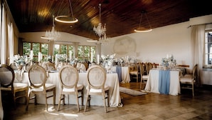 A spacious room with wooden ceilings, multiple tables set for dining, and large windows allowing natural light.