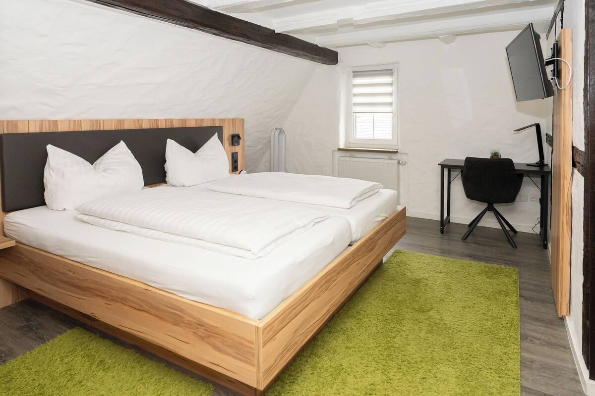 A bedroom with a wooden bed, white bedding, a green rug, a desk with a chair, and a TV mounted on the wall.