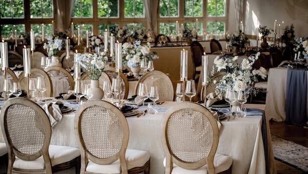 A formal dining room with tables set for multiple guests, featuring candle-lit centerpieces and wicker chairs.