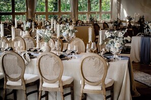 A formal dining room with tables set for multiple guests, featuring candle-lit centerpieces and wicker chairs.