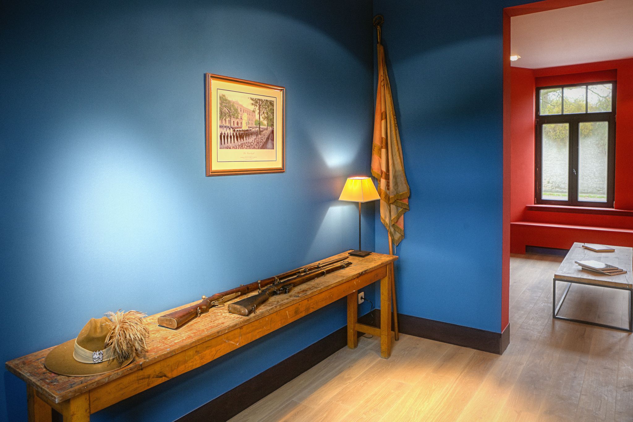 A room with a blue wall, a wooden bench with a hat and rifle, a framed picture, a lamp, and a window with a red frame.